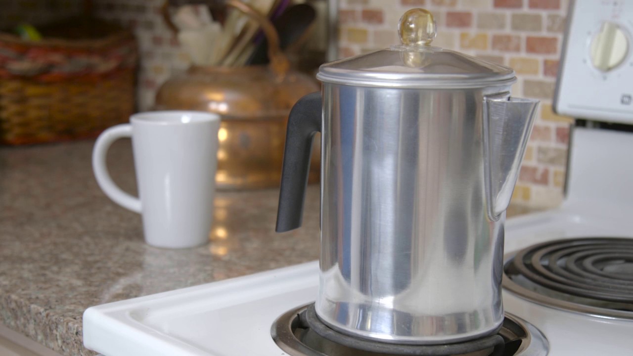 Metal percolator on stove top in front of white mug.